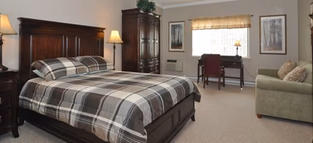 A bedroom with a large wooden bed featuring a plaid bedspread, two bedside tables with lamps, a wooden armoire, a desk with a chair near a window with a valance, and a green sofa with patterned pillows.