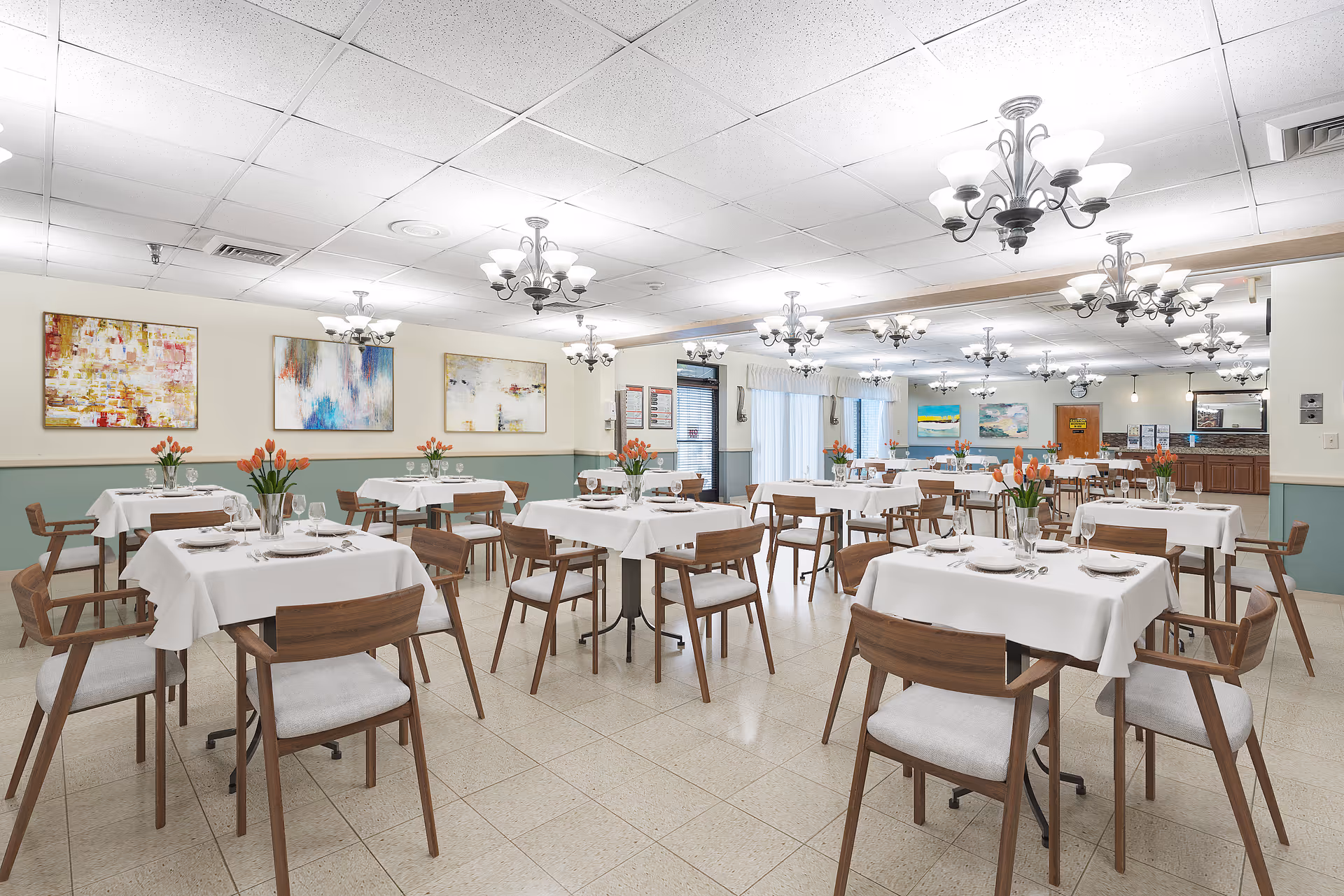 A spacious dining room with multiple tables covered in white tablecloths, each set with plates, glasses, and silverware. Wooden chairs with light cushions surround the tables. Vases with orange tulips decorate each table. The room is well-lit with multiple chandeliers hanging from the ceiling. Abstract paintings adorn the walls, and large windows with curtains allow natural light to enter.