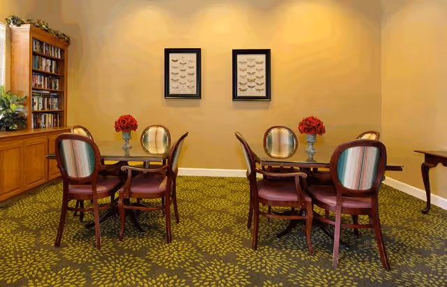 A cozy interior room with two wooden tables, each surrounded by four wooden chairs with striped upholstery. Each table has a vase with red flowers. On the left side, there is a wooden bookshelf filled with books and some greenery on top. The walls are painted a warm beige color with two framed artworks hanging in the center. The carpet has a green and yellow floral pattern.