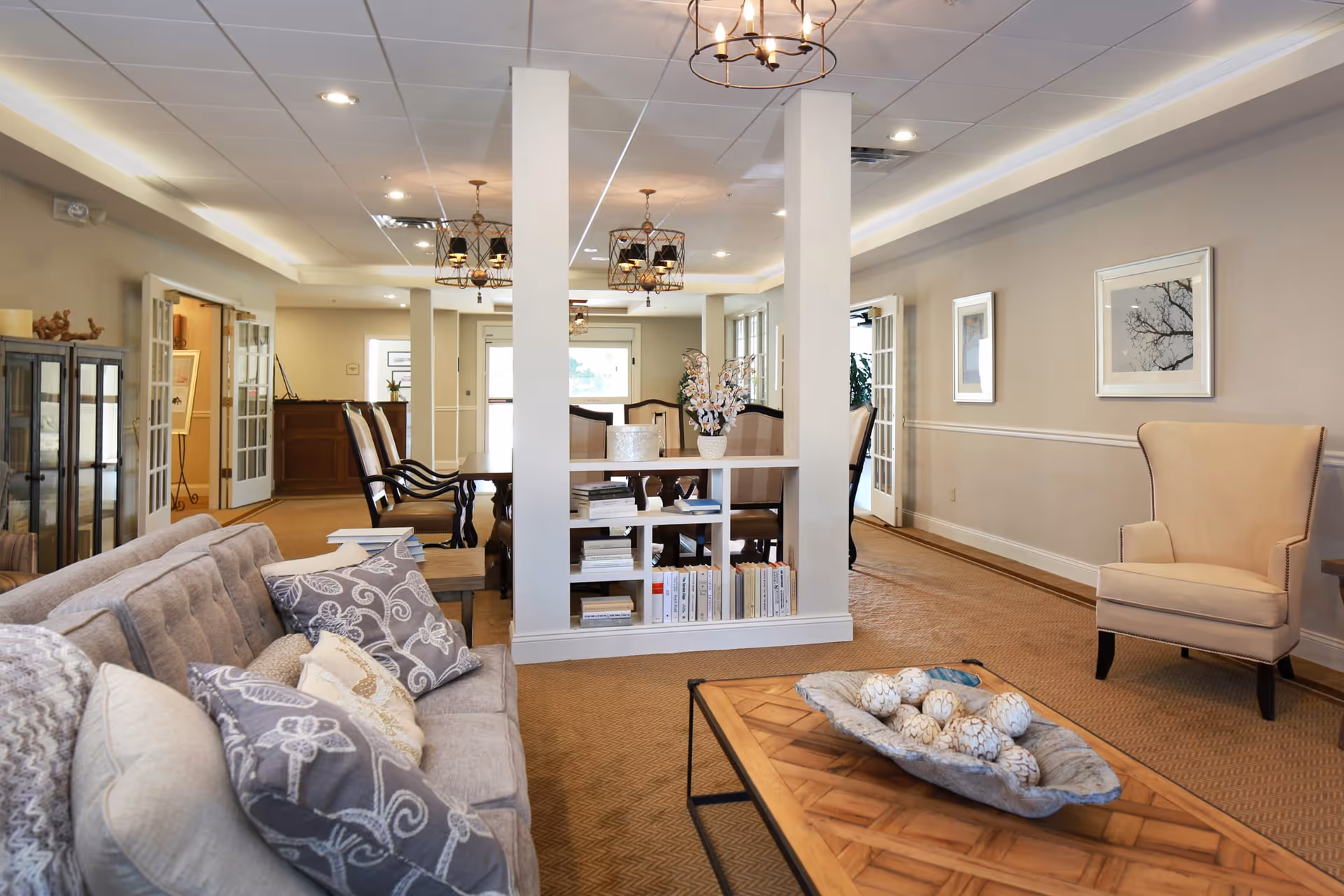 A cozy and well-lit living room area with a beige sofa adorned with patterned cushions, a wooden coffee table with decorative items, a beige armchair, and a white shelving unit with books and a flower vase. The room has beige walls with framed artwork, carpeted floors, and multiple hanging light fixtures. In the background, there is a dining area with a wooden dining table and chairs.