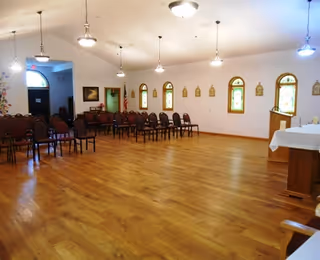 A spacious room with wooden flooring and multiple rows of brown chairs arranged along the walls. The room has several small arched windows with stained glass, pendant lights hanging from the ceiling, and a white table covered with a cloth on the right side. The walls are white, and there is a door and some framed pictures on the left side.