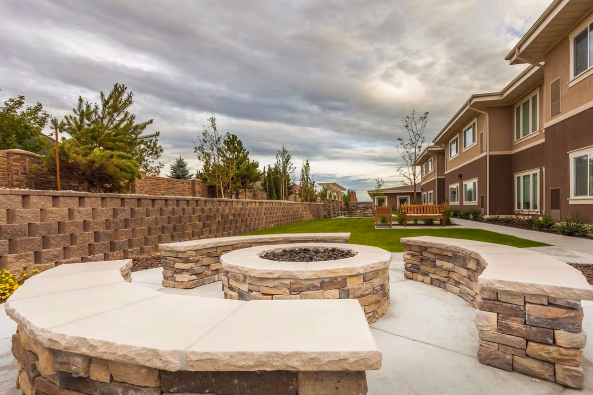 Outdoor courtyard with a circular stone fire pit and curved stone benches, lawn and seating beside a two-story building under a cloudy sky.