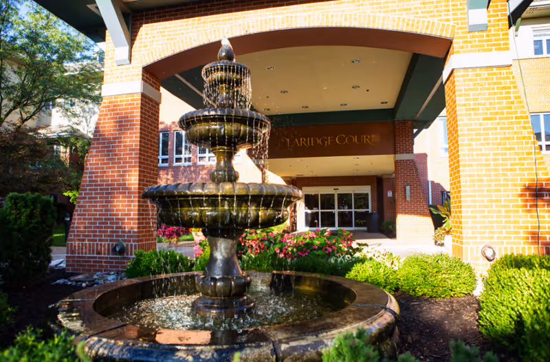 Outdoor view of the entrance to Claridge Court facility with a multi-tiered water fountain in the foreground surrounded by greenery and flowers, and a brick archway leading to the building entrance.
