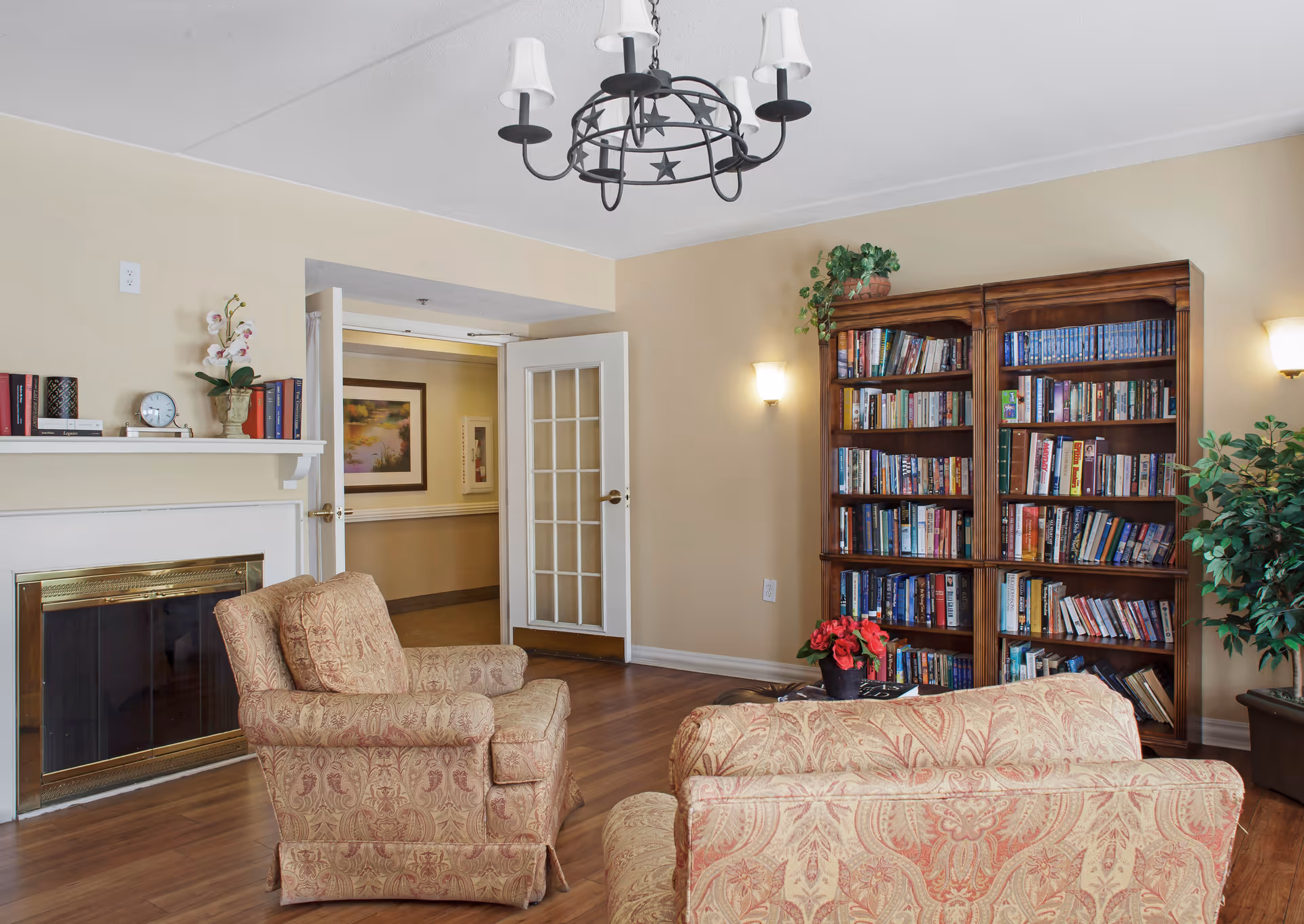 A cozy living room with two patterned armchairs and a matching sofa facing a fireplace with a white mantel. The mantel is decorated with books, a clock, and a flower vase. There is a large wooden bookshelf filled with books against the wall, with a potted plant on top. The room has wooden flooring, beige walls, a chandelier with white lampshades, and wall sconces providing warm lighting. A door with glass panels is open, showing a hallway with framed artwork.