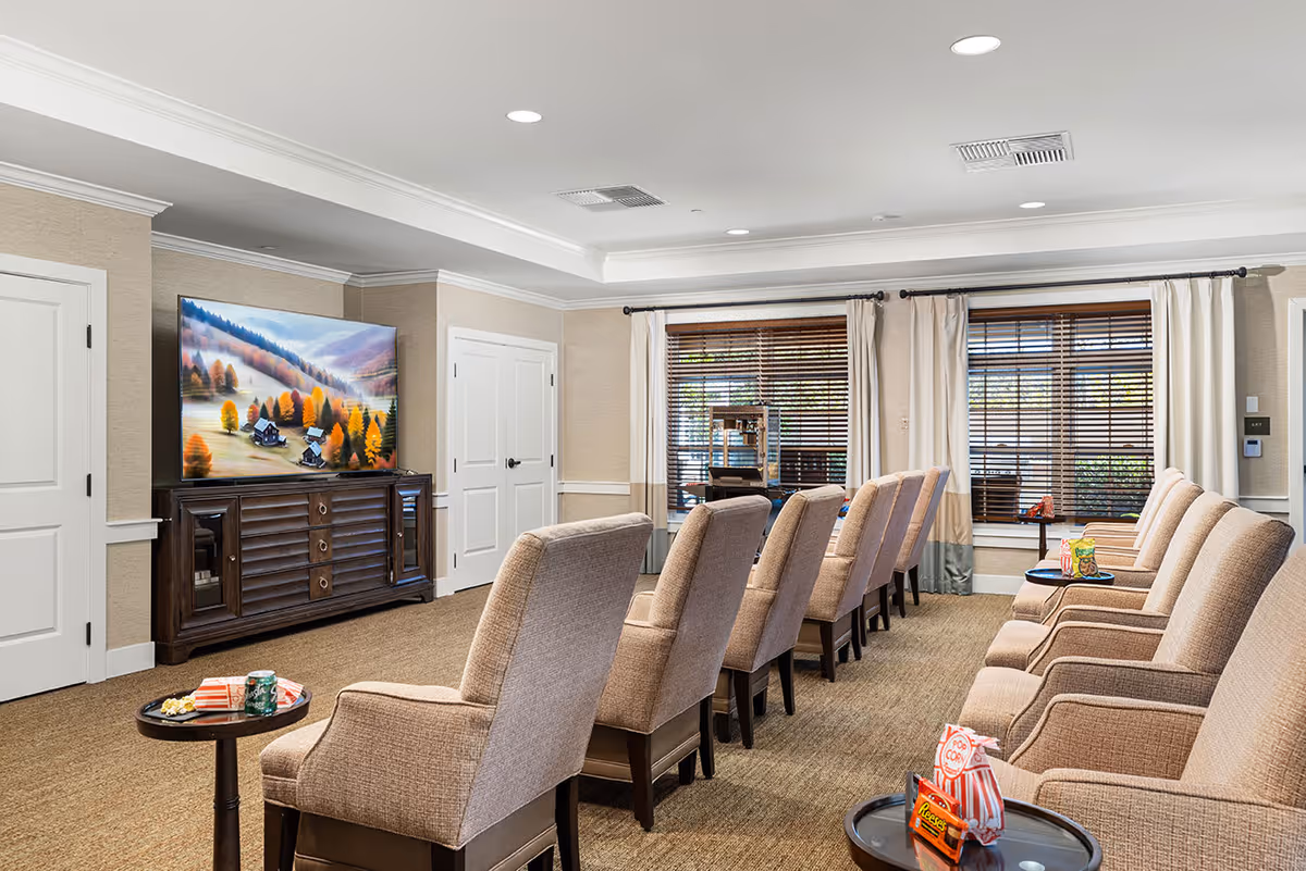 A cozy living room with two rows of beige upholstered armchairs facing a large flat-screen TV mounted on a wooden cabinet. Small round tables beside the chairs hold snacks like popcorn and candy. The room has beige walls, carpeted floor, and large windows with wooden blinds and cream-colored curtains.