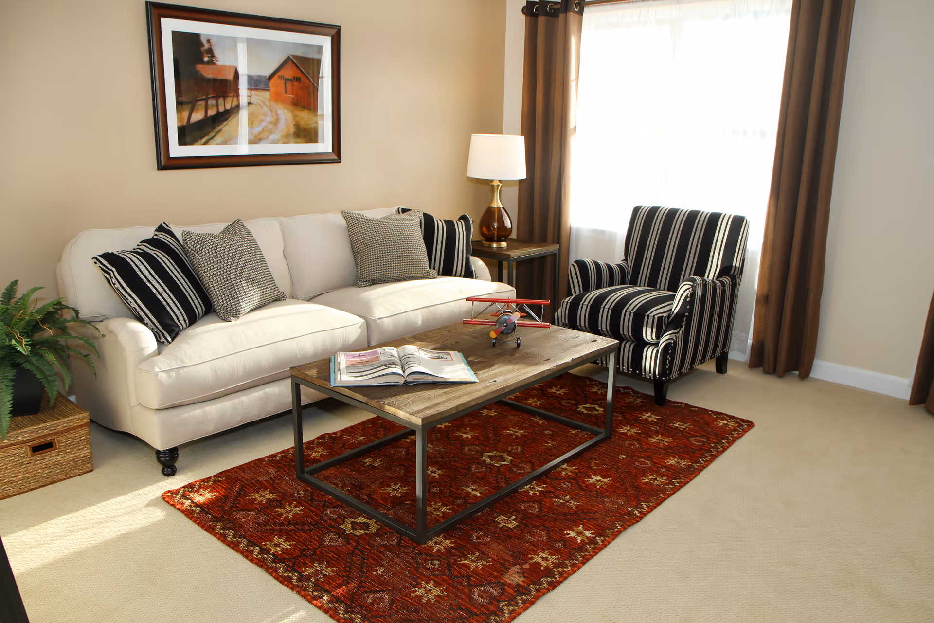 A cozy living room with a white sofa adorned with black and white striped and patterned pillows, a wooden coffee table with an open book and a small model airplane on it, a striped armchair, a side table with a lamp, a red patterned rug, a framed painting of a barn on the wall, and a window with brown curtains letting in natural light.
