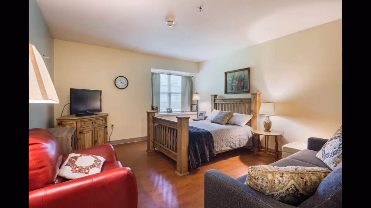 A cozy bedroom in a senior living facility featuring a wooden bed with pillows and a blanket, a red armchair with a decorative pillow, a gray sofa with patterned cushions, a wooden dresser with a flat-screen TV, a wall clock, a window with curtains, and two table lamps providing warm lighting.