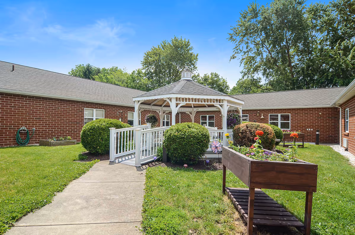 Outdoor courtyard area at Celebration Villa of Berwick featuring a white gazebo with a ramp, surrounded by green bushes, a flower planter with colorful flowers, and a brick building in the background under a clear blue sky.