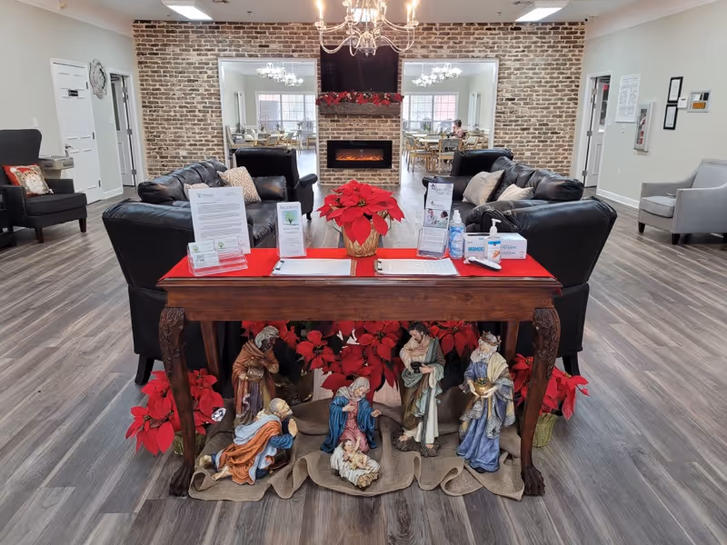 A cozy senior living community common area with black leather sofas arranged around a modern electric fireplace set in a brick wall. A wooden table in the foreground is decorated with red poinsettias and a nativity scene. The room has wood flooring, light-colored walls, and a chandelier hanging from the ceiling. In the background, a dining area with tables and chairs is visible through two large openings in the brick wall.