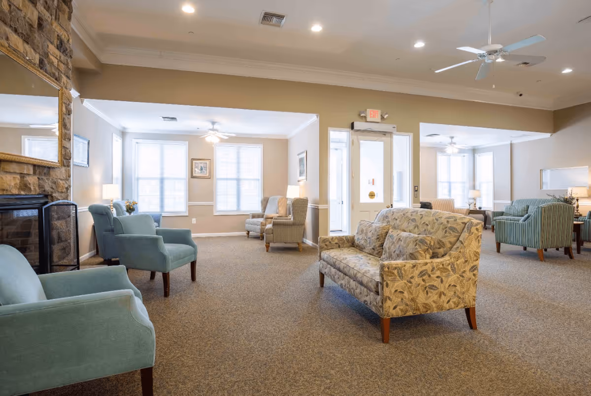 A spacious senior living facility common area with multiple upholstered armchairs and sofas arranged around the room. The room features beige walls, carpeted floors, ceiling fans, and large windows letting in natural light. A stone fireplace with a large mirror above it is visible on the left side.