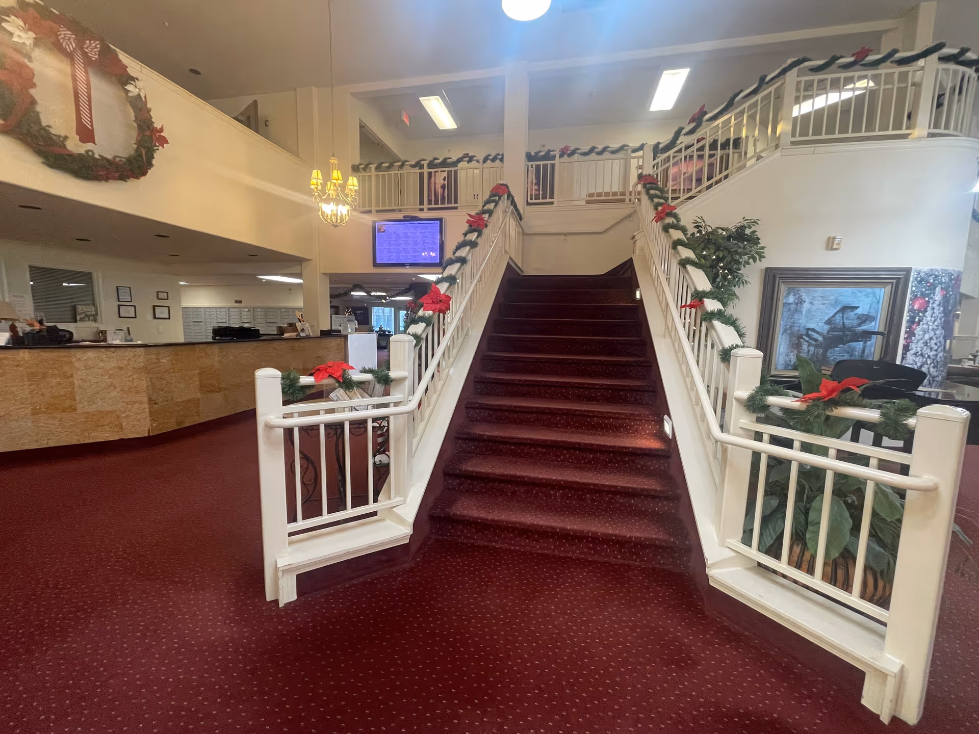 Interior view of a senior living facility lobby with a red carpeted staircase decorated with garlands and red bows. To the left is a reception desk with a granite front, and above it hangs a chandelier. The upper level has white railings also decorated with garlands and red bows. There is a large wreath on the wall to the left and a framed painting on the right side near some plants.
