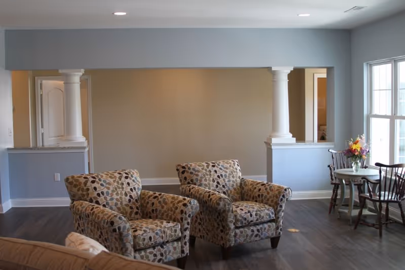 A cozy living area with two patterned armchairs facing forward, a small round table with a flower vase and two wooden chairs near large windows letting in natural light. The room has light blue and beige walls, white columns, and dark wood flooring.