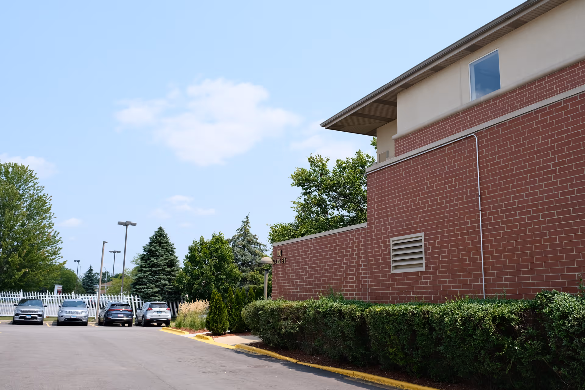 Exterior view of a brick building with a parking lot in front, several parked cars, green bushes along the building, and trees in the background under a partly cloudy sky.