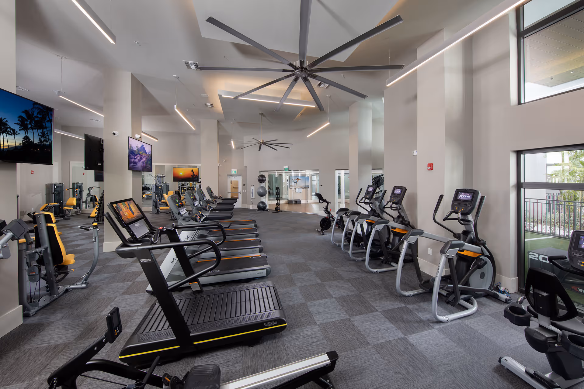 A modern fitness center with multiple treadmills, elliptical machines, and exercise bikes arranged in rows. The room has large windows letting in natural light, high ceilings with large ceiling fans, and several wall-mounted televisions. The flooring is carpeted with a checkered pattern, and there are exercise balls and other gym equipment visible in the background.
