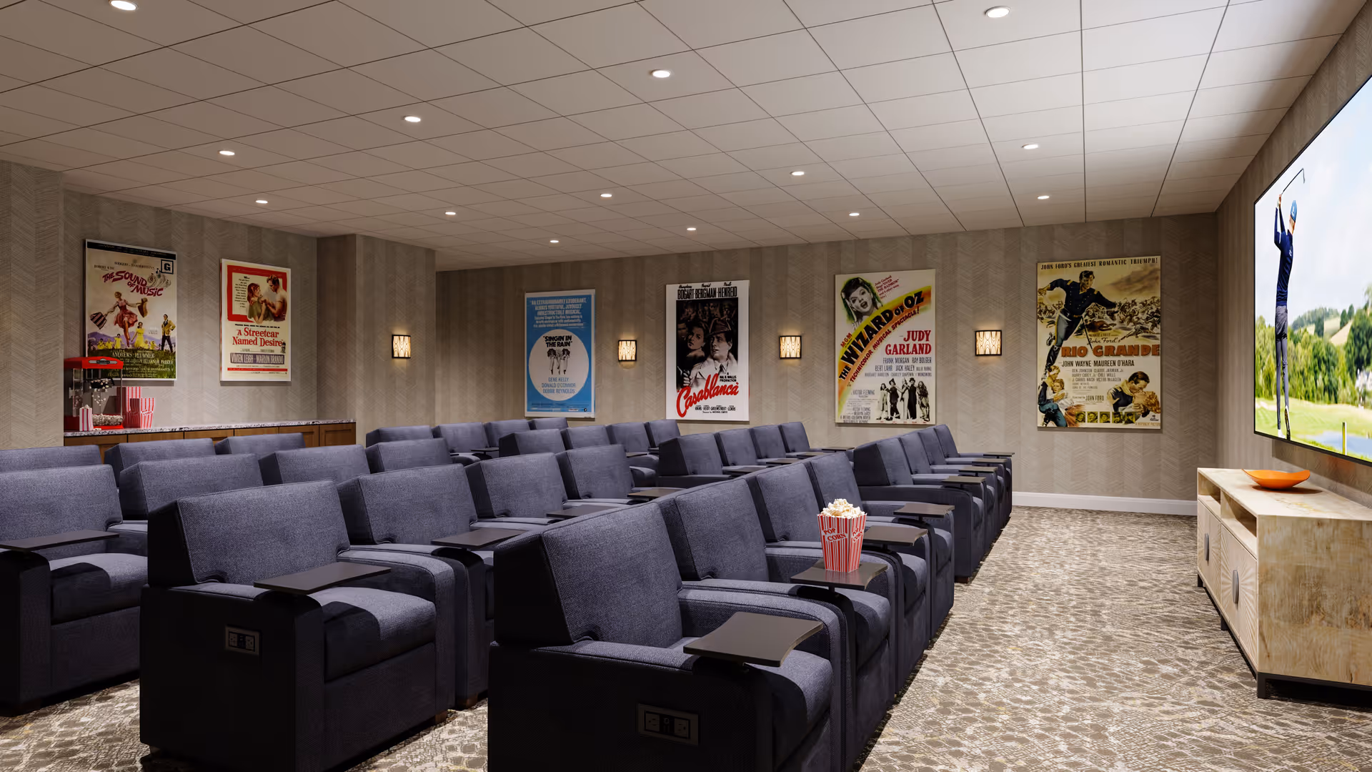 A cozy movie theater room with multiple rows of dark gray reclining seats, each equipped with a small black tray table. A large flat-screen TV is mounted on the right wall, displaying a golf scene. Classic movie posters decorate the back wall, and a popcorn machine is visible on a counter to the left. The room has a patterned carpet and recessed ceiling lights.