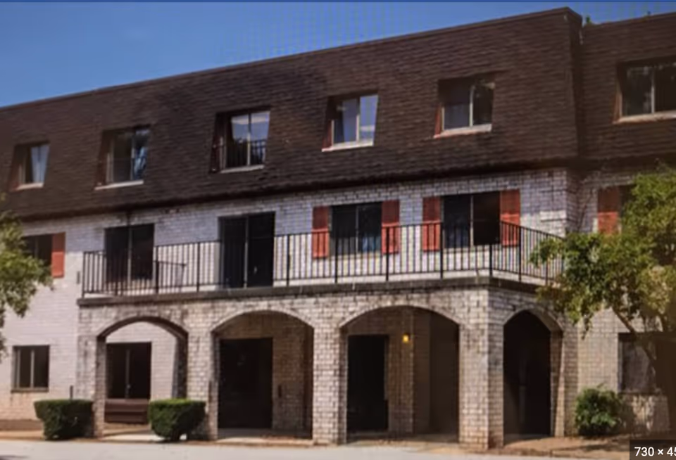 Exterior view of a multi-story brick building with arched entrances on the ground floor, a balcony with black railing on the second floor, and several windows with red shutters. There are small bushes and trees near the building under a clear blue sky.