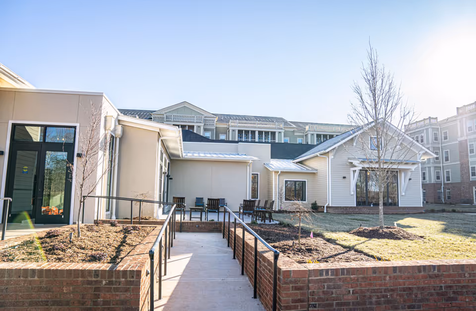 Outdoor view of The Woodlands at Furman facility showing a paved walkway with handrails leading to a patio area with chairs. The building exterior is light-colored with large windows, and there are small trees and landscaped garden beds along the walkway. The sky is clear and sunny.