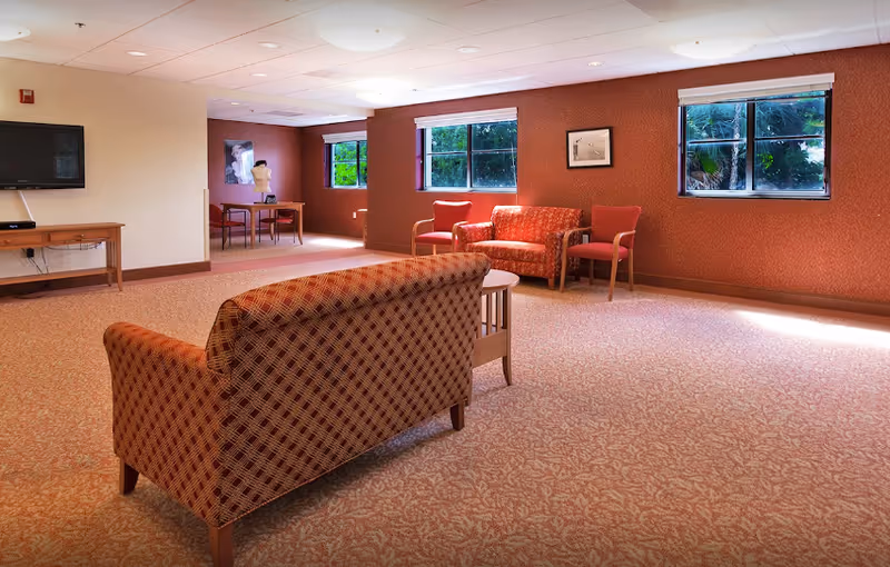 Spacious communal lounge with patterned sofas and chairs, side tables, a wall-mounted TV, and windows overlooking greenery.