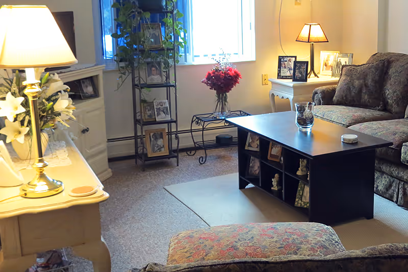 A cozy living room with a floral patterned sofa and matching ottoman, a black coffee table with decorative items, a white side table with a lamp and framed photos, a tall plant stand with more framed photos and a plant, and a window letting in natural light.