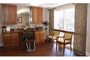 A well-lit room with wooden flooring featuring a barber chair in front of a counter with cabinets and a mirror. Next to the chair, there are two wooden chairs with cushions placed near a large window with patterned curtains.