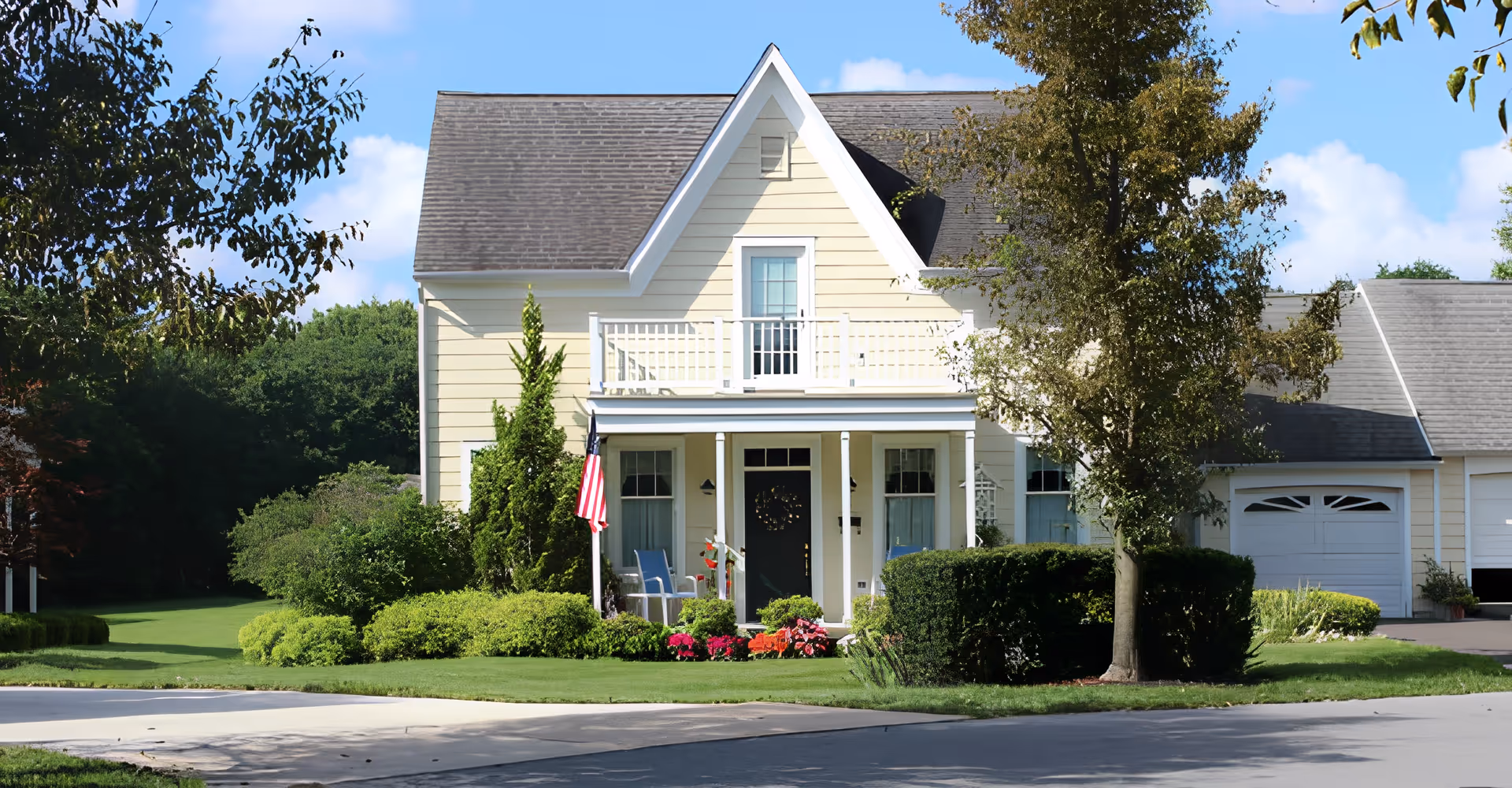 Front exterior view of a two-story yellow house with a gray roof, a small balcony above the porch, an American flag, and well-maintained green bushes and flowers in the yard.