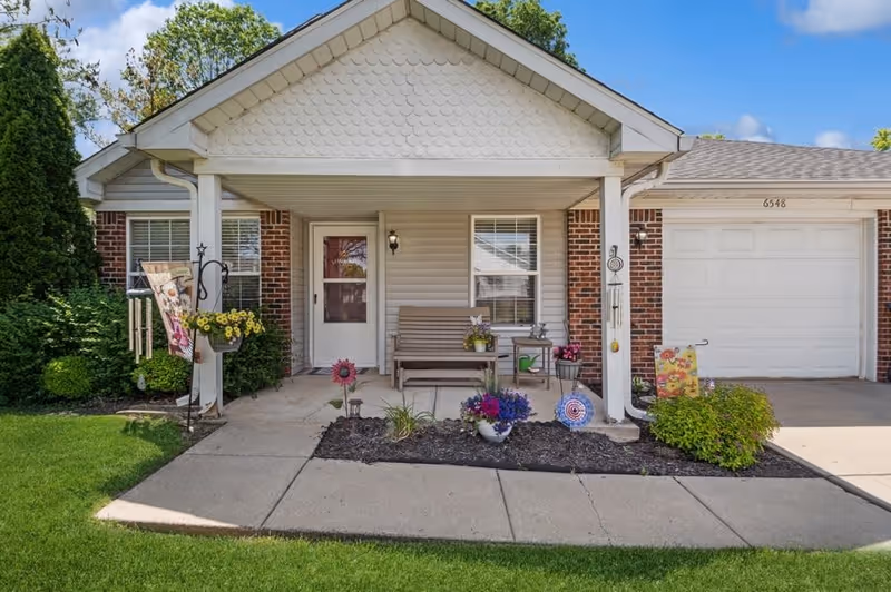 Front porch of a single-story residence with a covered entryway, a bench, potted flowers, decorative garden flags, and a garage door to the right. The house exterior features brick and siding with a small garden bed in front.