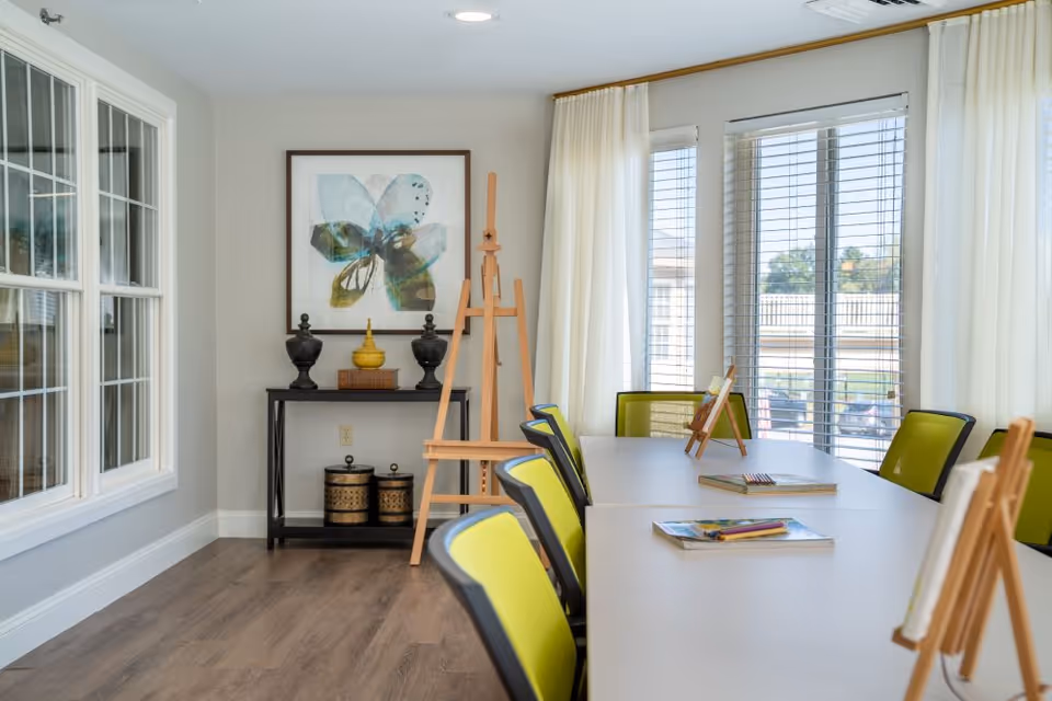 A bright room with large windows covered by white curtains and blinds, featuring a long table with green and black chairs around it. On the table are small wooden easels holding canvases and some coloring books with colored pencils. Against the wall is a small black console table with decorative items and a framed abstract flower artwork above it. The floor is wooden and the room has a clean, modern look.