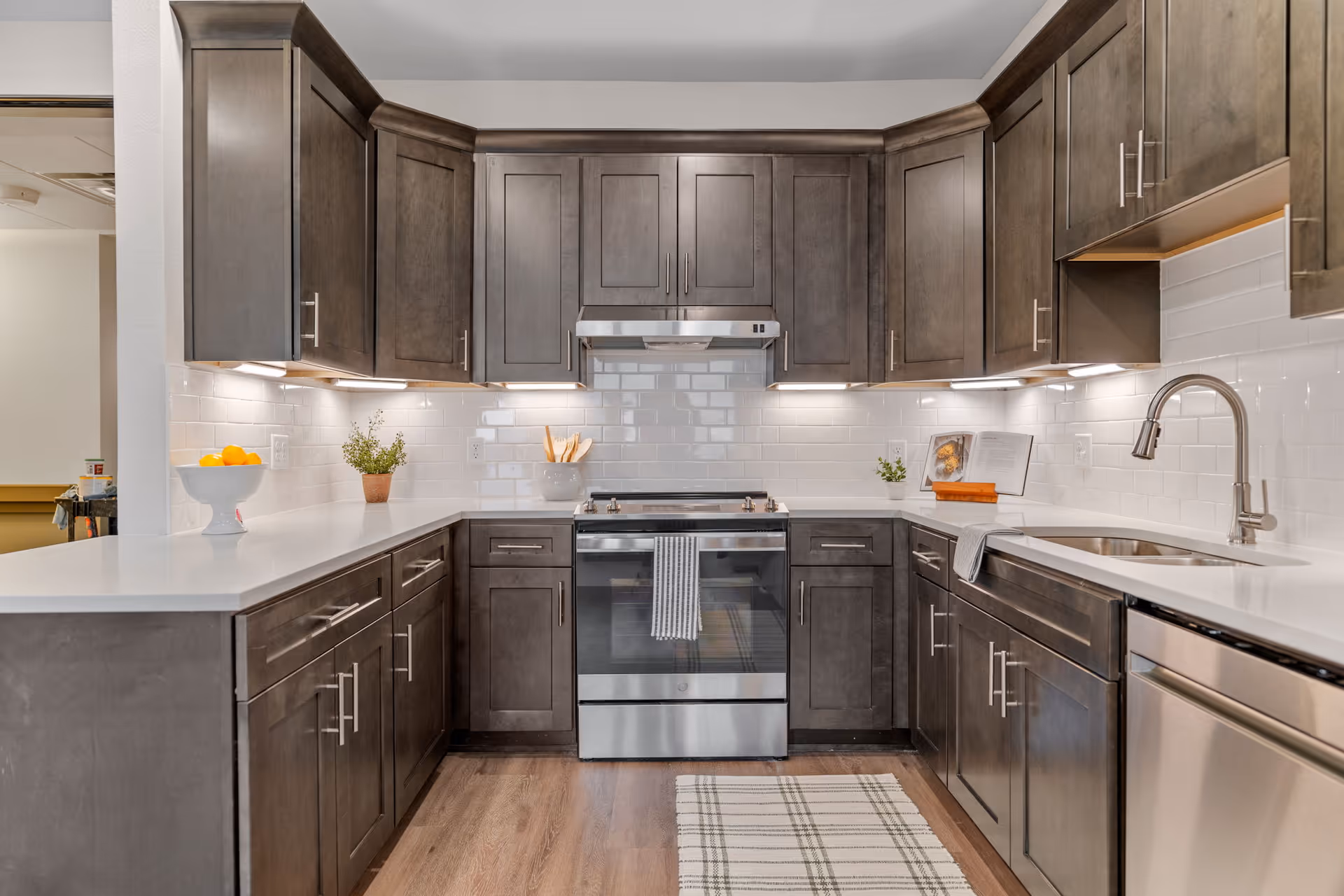 Modern kitchen with dark wood cabinets, white countertops, stainless steel oven and dishwasher, white subway tile backsplash, under-cabinet lighting, a sink with a curved faucet, and a striped rug on the wooden floor.