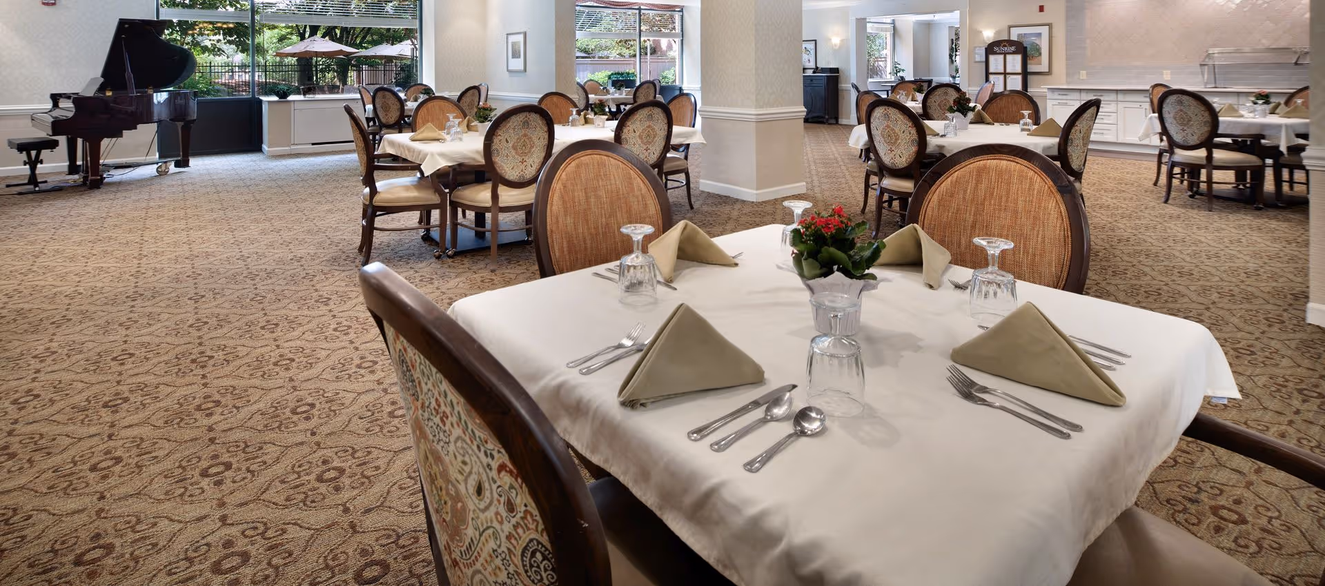 Well-lit dining room with tables set with napkins and glassware, upholstered chairs, a grand piano, and large windows.
