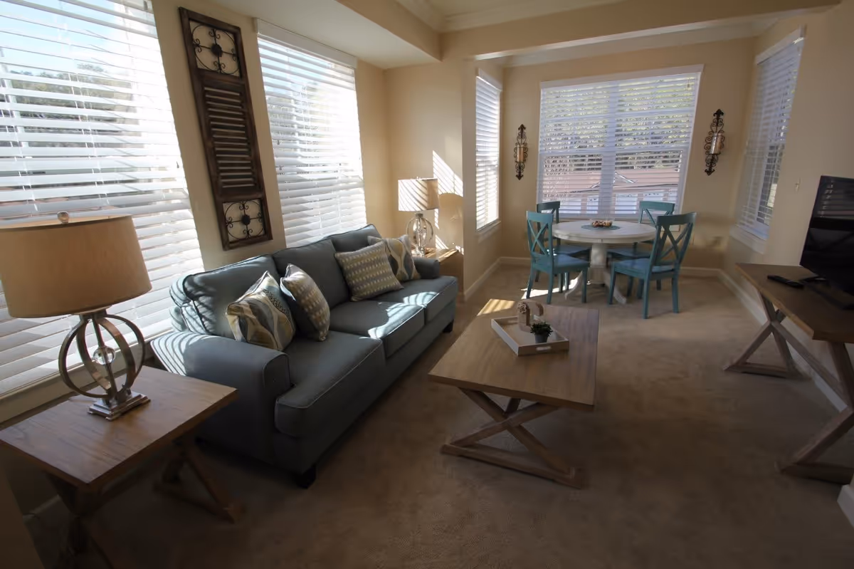 A cozy living room with a blue sofa adorned with patterned cushions, a wooden coffee table with a small plant and tray, a side table with a lamp, and a round dining table with four blue chairs near large windows with white blinds letting in natural light.