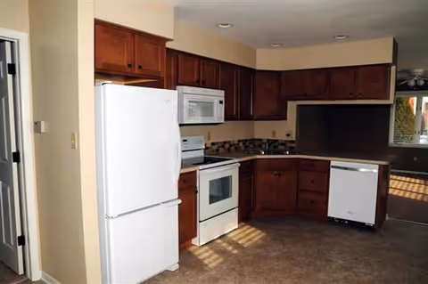 Open kitchen with white refrigerator, stove, microwave and dishwasher surrounded by dark wood cabinets.