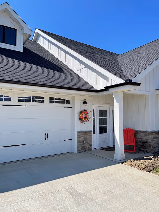 Exterior view of a white house with a black shingled roof, a white garage door with decorative handles, a front door with glass panels, a colorful wreath hanging beside the door, a red Adirondack chair, and a small black side table on the porch under a clear blue sky.