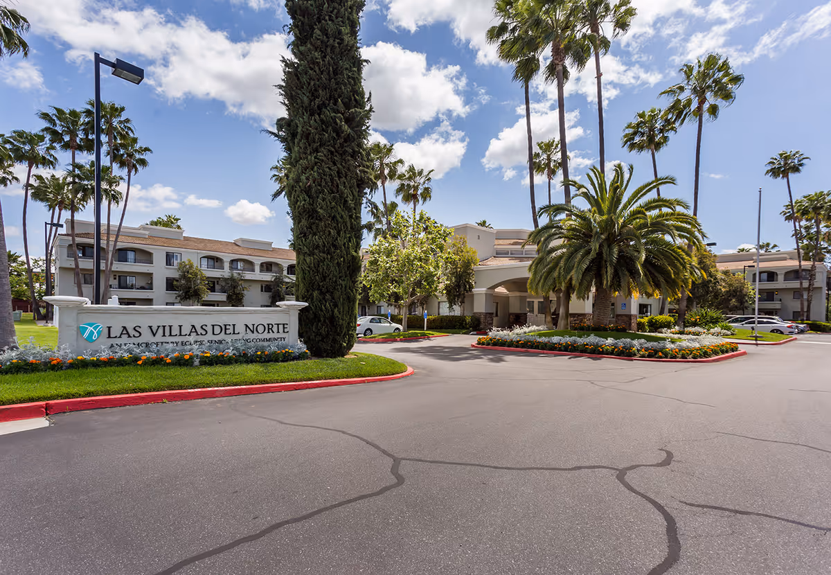 Exterior view of Las Villas Del Norte senior living community with a driveway, landscaped flower beds, palm trees, and a multi-story building in the background under a partly cloudy sky.