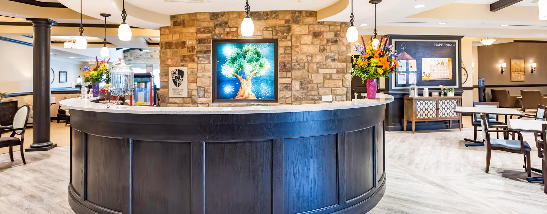 A spacious and well-lit common area in a senior living facility featuring a curved dark wood reception desk with a white countertop. Behind the desk is a stone accent wall with a colorful framed artwork of a tree. The room includes tables and chairs, a bulletin board labeled 'Happenings,' and floral arrangements on the desk and a sideboard.