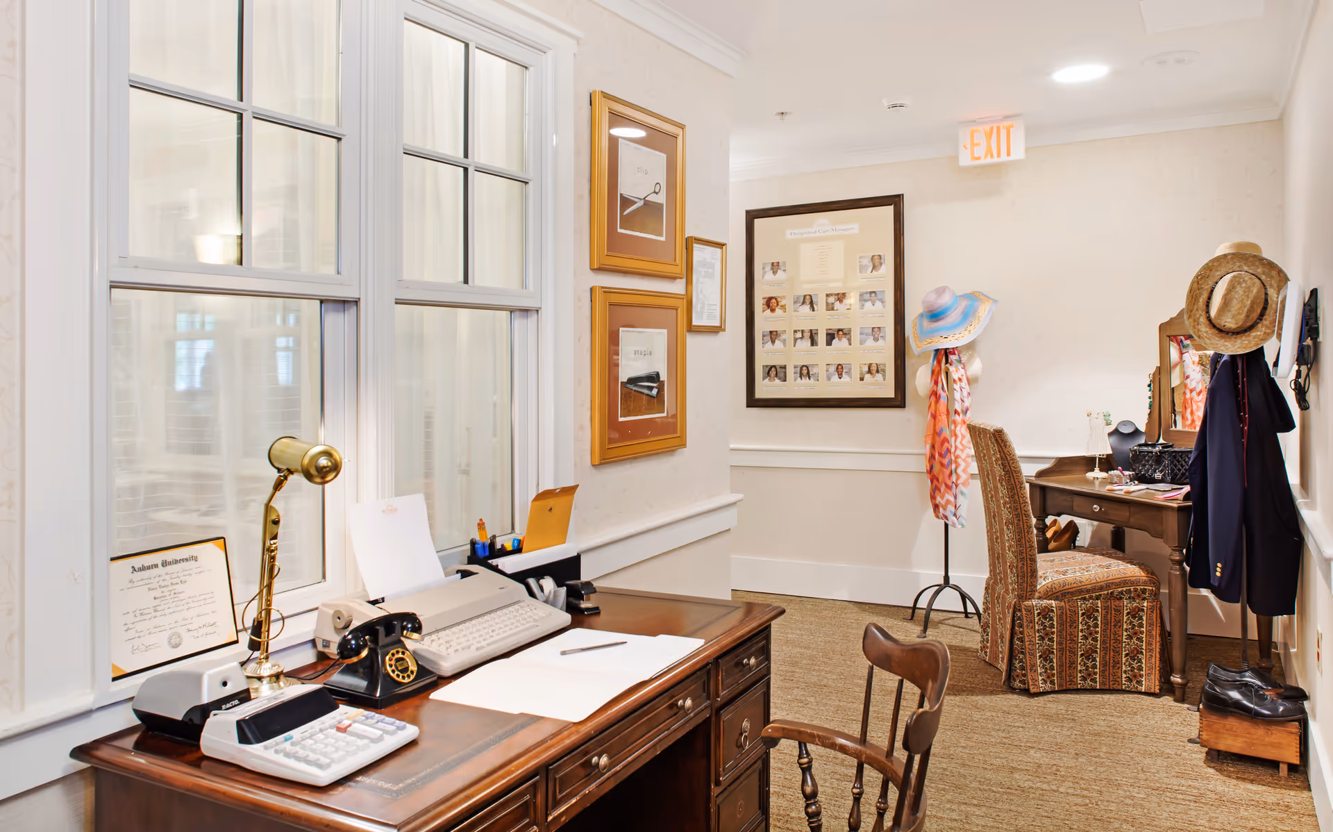 Well-lit interior hallway with wooden desks and chairs, vintage office equipment on a desk, and a small dressing area with hats and a mannequin.