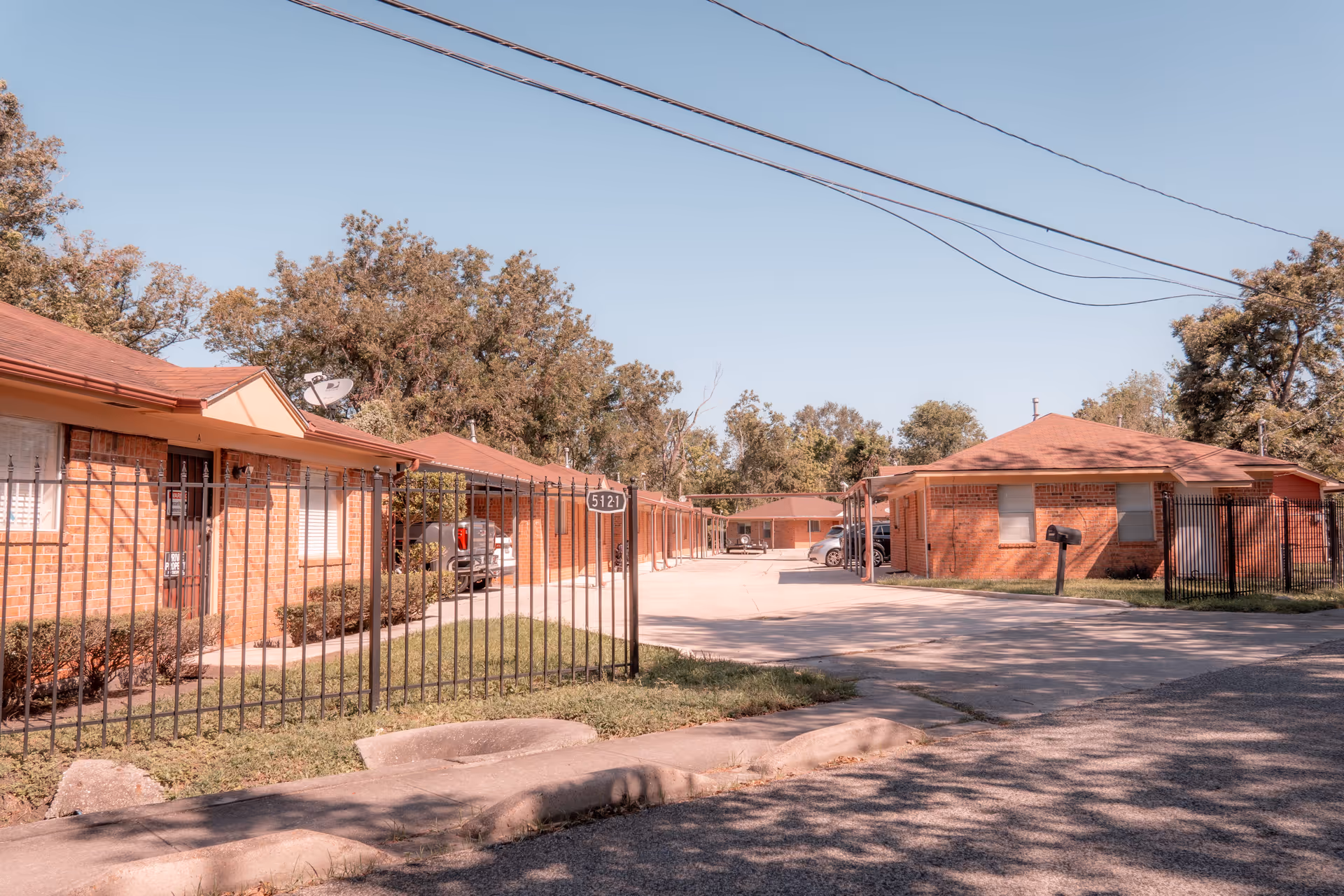 Exterior view of a single-story brick assisted living facility with multiple units, a gated entrance, parked cars under carports, and trees in the background under a clear blue sky.