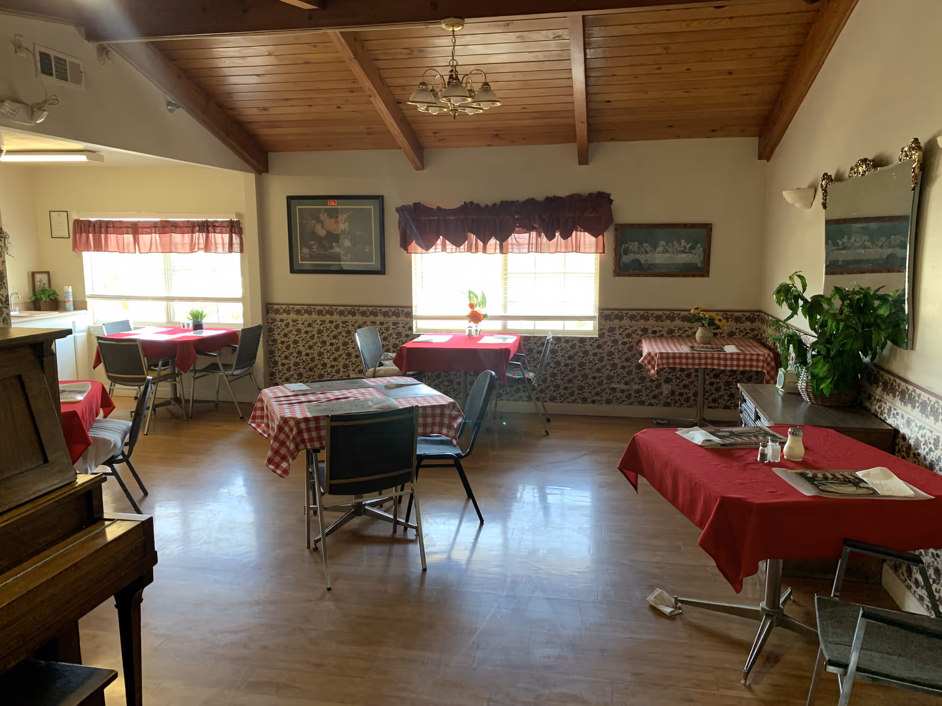 A cozy dining room with wooden ceiling beams and several tables covered with red and red-checkered tablecloths. Each table has chairs around it, placemats, and some have small plants or flowers as centerpieces. The room has large windows with red valances letting in natural light, floral wallpaper halfway up the walls, and framed artwork. There is a piano on the left side and a large mirror on the right wall above a sideboard with a green plant.