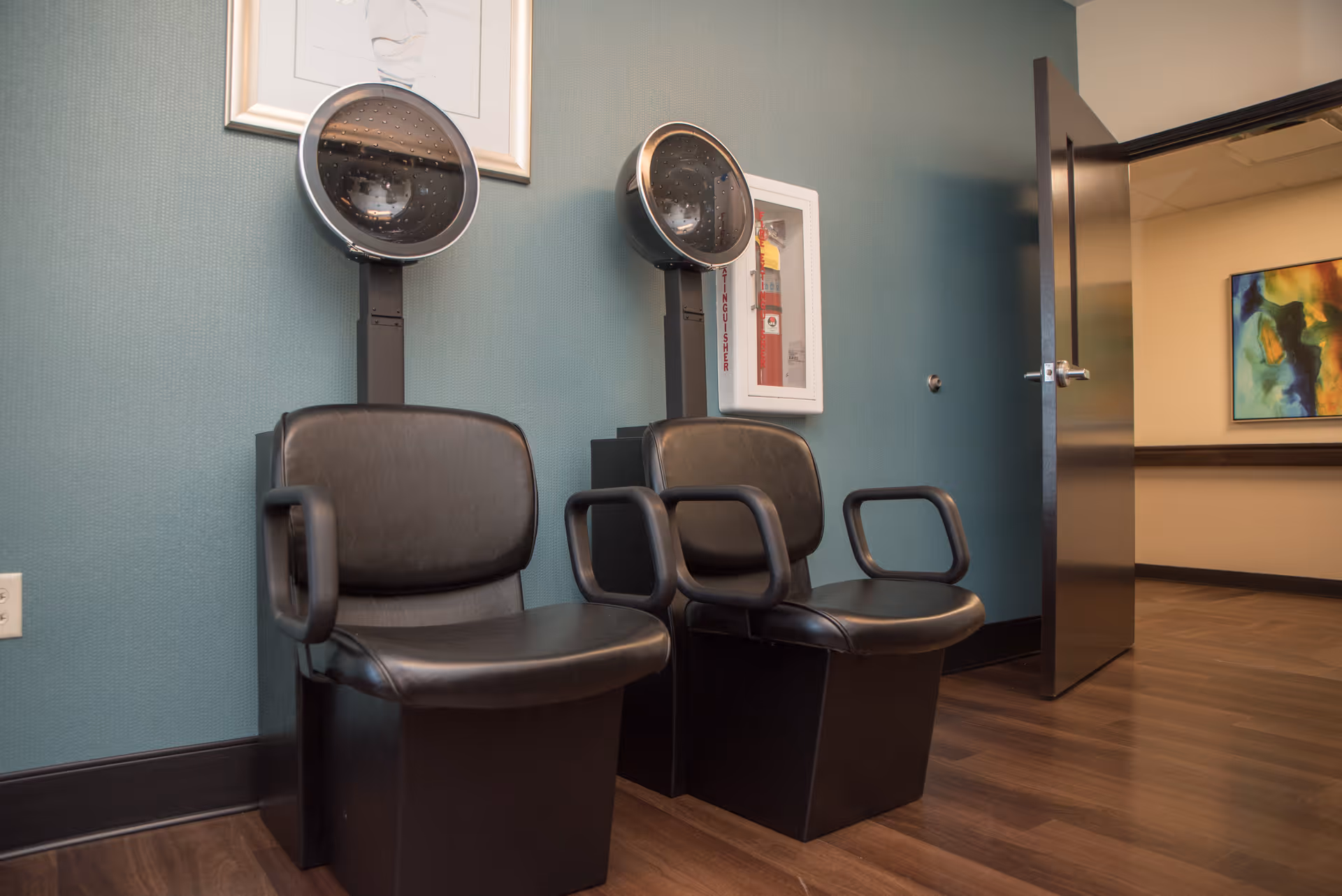 Two black salon hair dryer chairs with overhead hair drying hoods against a teal wall, next to an open door leading to a hallway with a colorful abstract painting on the wall.