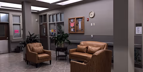 Interior lounge with brown leather seating, plants, bulletin board, and clock on gray walls.