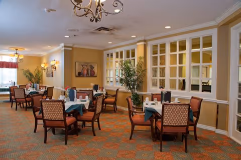 Warm dining room with several set tables, upholstered chairs, chandeliers, and potted plants.