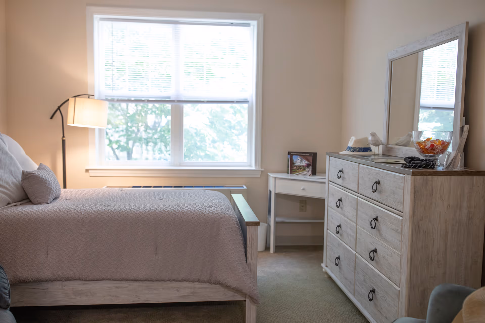 A bedroom with a large window letting in natural light, a bed with light-colored bedding and pillows, a floor lamp beside the bed, a white desk with a framed picture on it, and a wooden dresser with a mirror, decorative items, and a bowl of candy on top.
