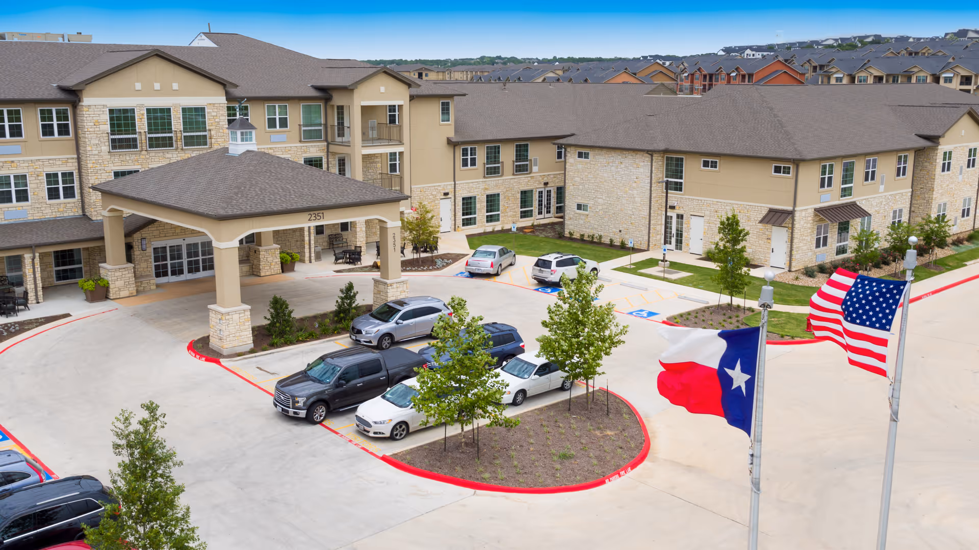 Exterior view of The Enclave at Round Rock Senior Living facility showing a large building with beige and stone facade, multiple windows, and a covered entrance. Several cars are parked in the driveway and parking lot. Two flagpoles display the Texas state flag and the United States flag. The surrounding area includes small trees and landscaped greenery under a clear blue sky.
