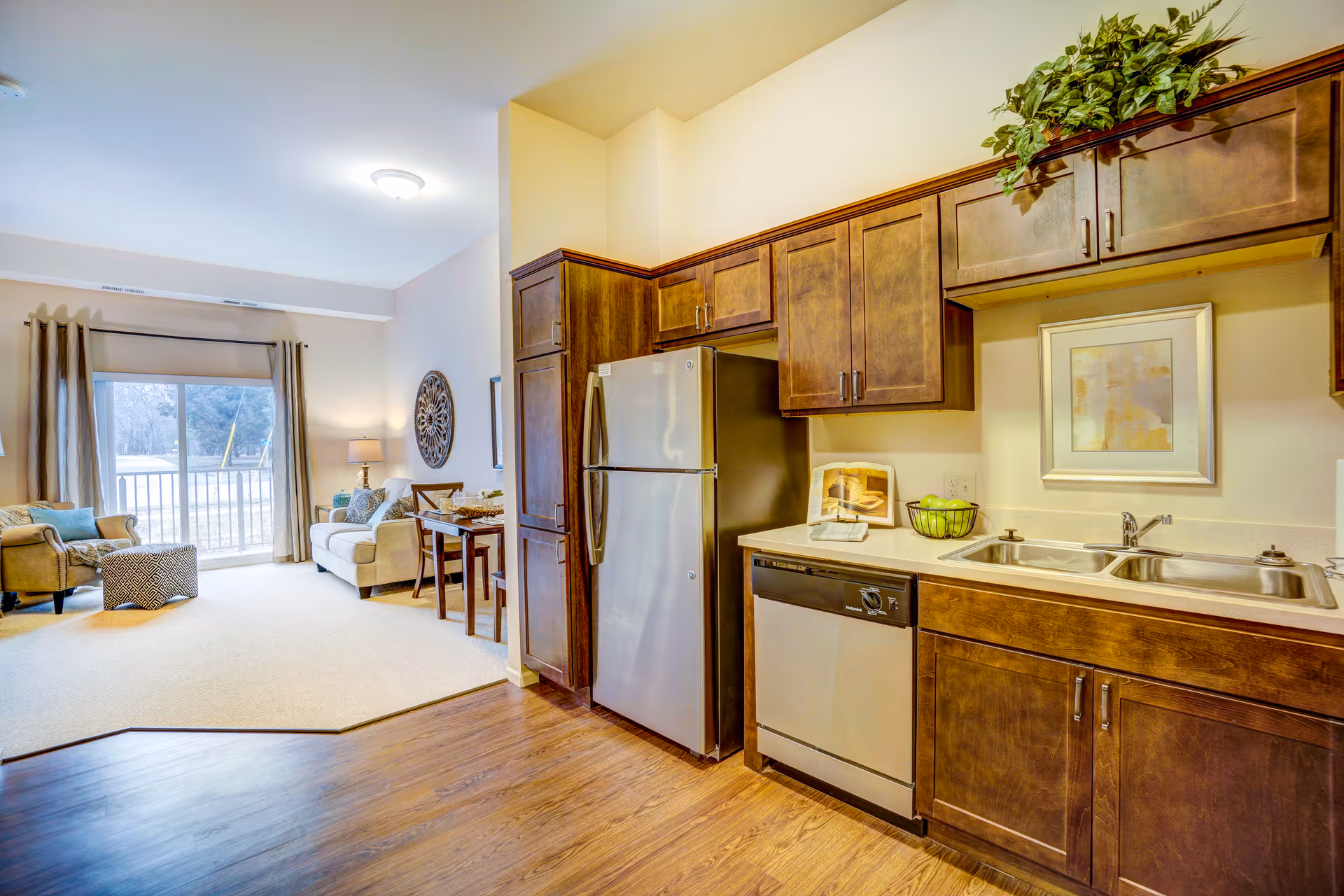 Interior view of a senior living facility apartment at Gable Pines showing a kitchen area with wooden cabinets, a stainless steel refrigerator, dishwasher, and double sink. Adjacent to the kitchen is a living area with a beige sofa, armchair, small dining table with chairs, and a large window with curtains letting in natural light.