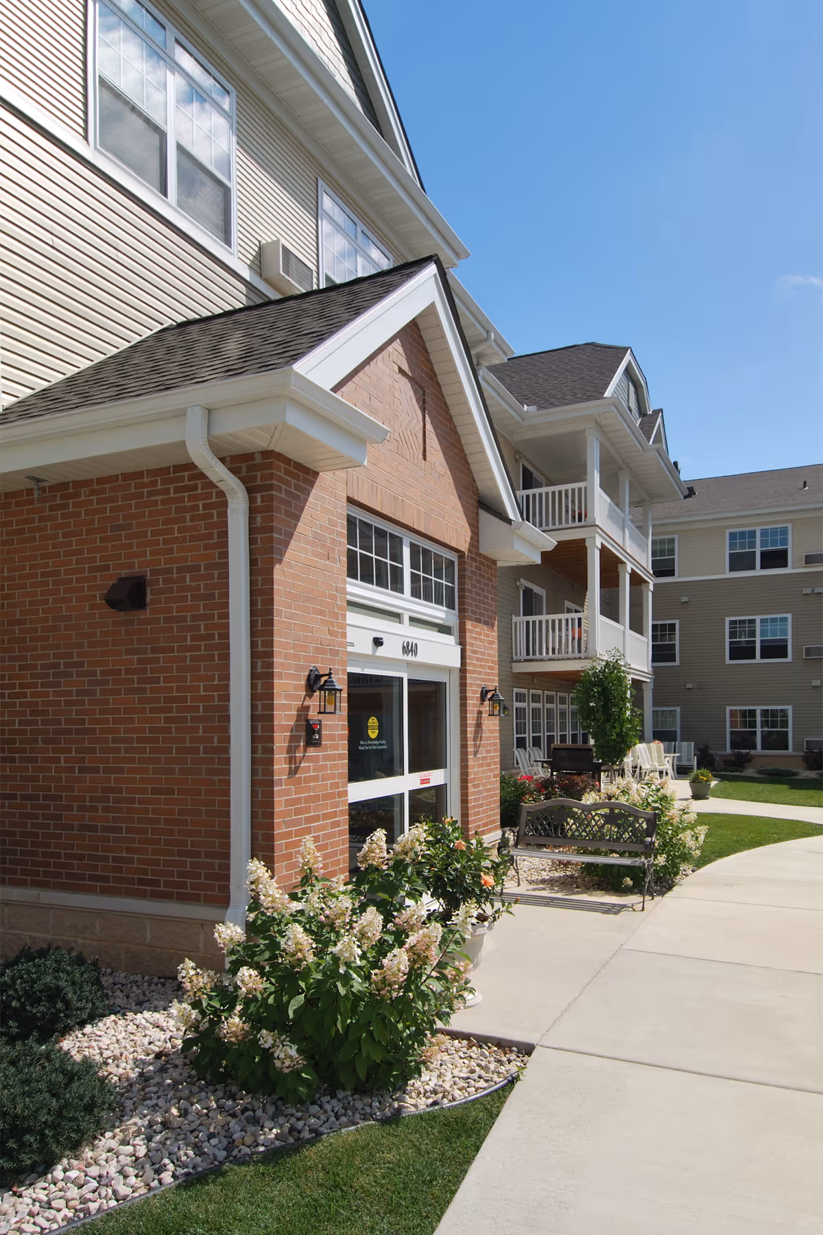Front entrance of a multi-story senior apartment building with a brick facade, balconies, landscaped walkway and bench.