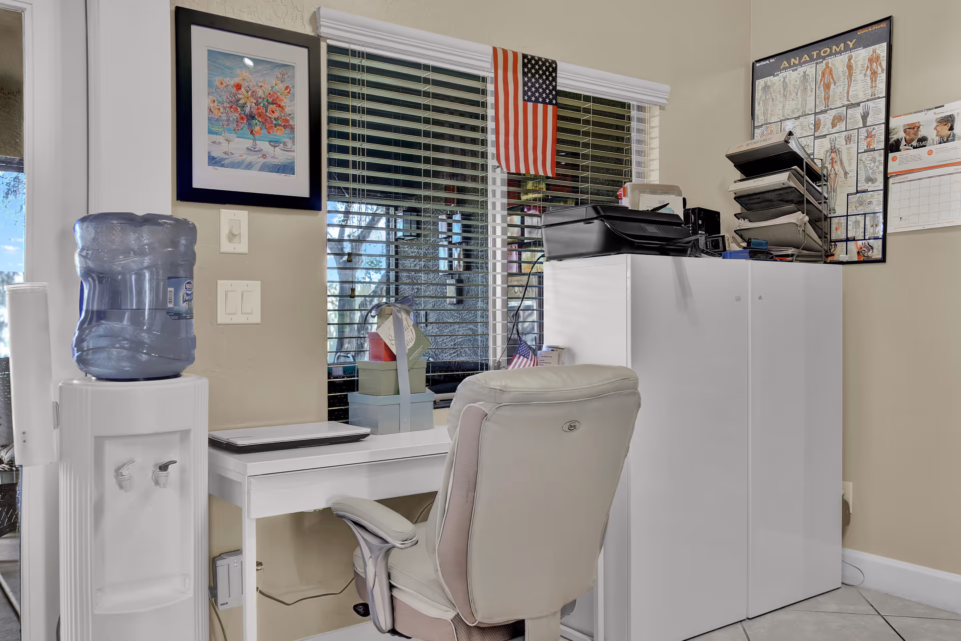 A small office area with a white desk and beige office chair facing a window with blinds. On the desk are a closed laptop and some gift boxes. To the left is a water cooler with a large water jug. On the right side, there is a white cabinet with a printer and office supplies on top. The walls have a framed floral painting, an anatomy chart, and a calendar. An American flag is hanging in the window.