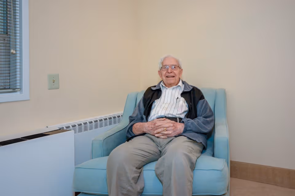 An elderly man with white hair and glasses sitting and smiling on a light blue armchair in a room with beige walls and a window with blinds.