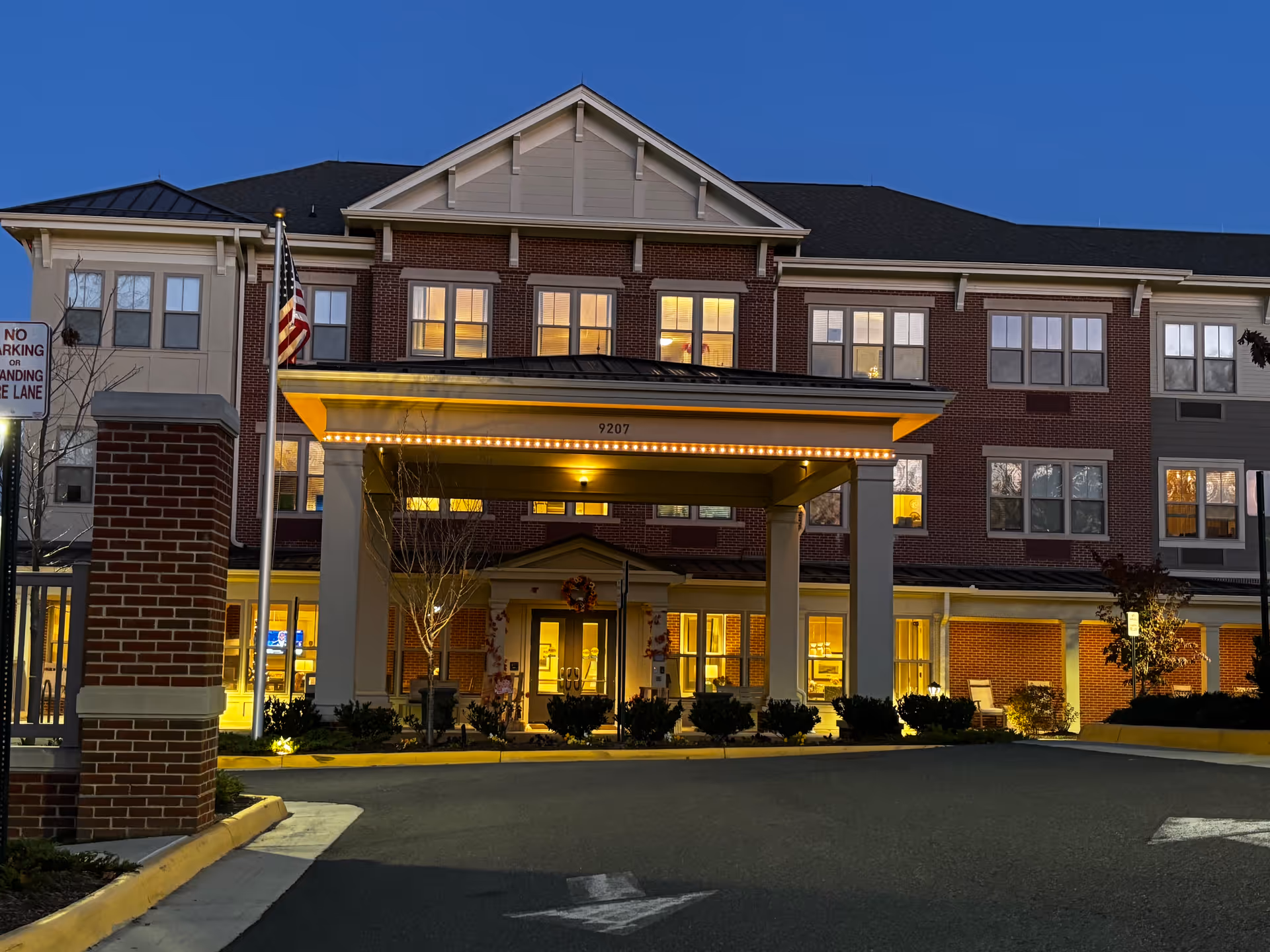 Exterior front view of Sunrise of Fairfax senior living facility at dusk, showing a three-story brick building with illuminated windows, a covered entrance with lights, an American flag on a flagpole, and a 'No Parking or Standing Fire Lane' sign.