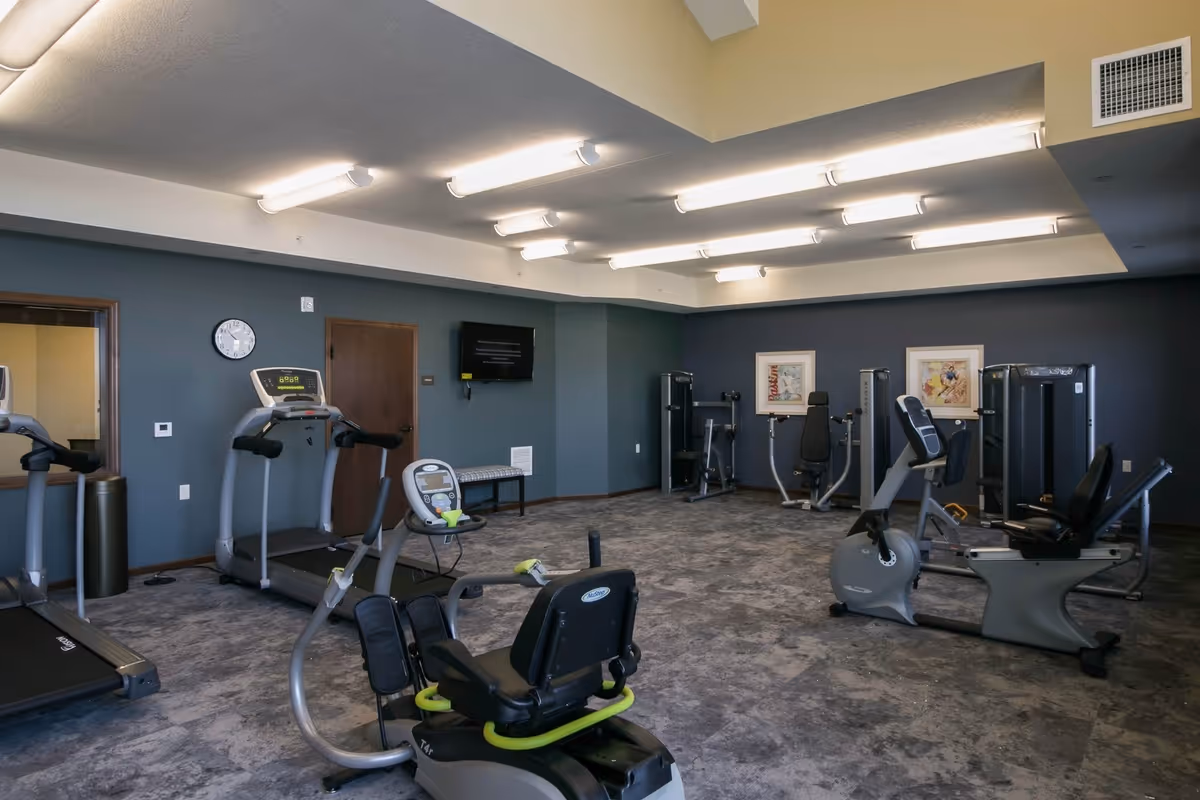 A fitness room with various exercise equipment including treadmills, stationary bikes, and weight machines. The room has gray walls, a clock on the wall, a mounted TV, and framed artwork. The ceiling has multiple fluorescent lights.
