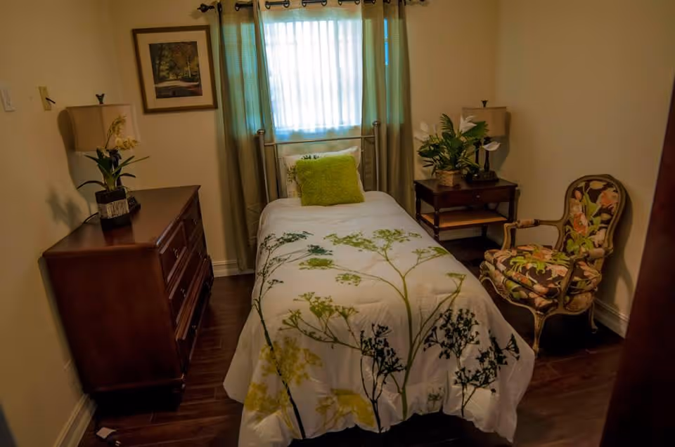 A small bedroom with a single bed covered in a white comforter with green and yellow floral patterns. There is a green pillow on the bed. The room has wooden flooring, a wooden dresser with a lamp and a plant on it, a side table with a lamp and a plant, a floral upholstered armchair, and a window with sheer curtains letting in natural light.