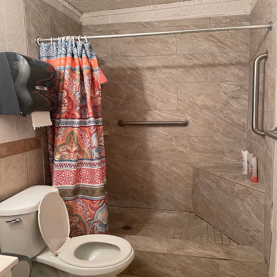 Tiled accessible bathroom with a toilet (lid open), a colorful patterned shower curtain, a roll-in shower with corner bench and stainless grab bars.
